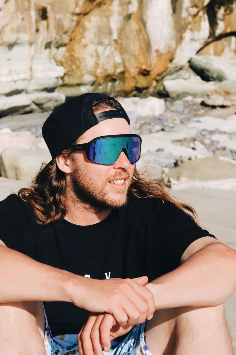 A man in wraparound CARVE sport sunglasses and a cap, sitting on the rocks