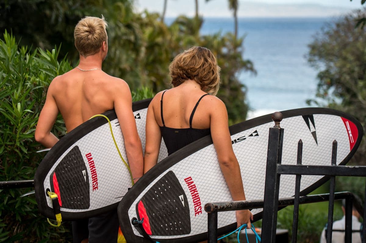 Two riders from behind, carrying boards toward the water